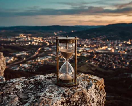 An hourglass on a hilltop at dawn, cupped by a hand of light, the cities of the nations in the valley below